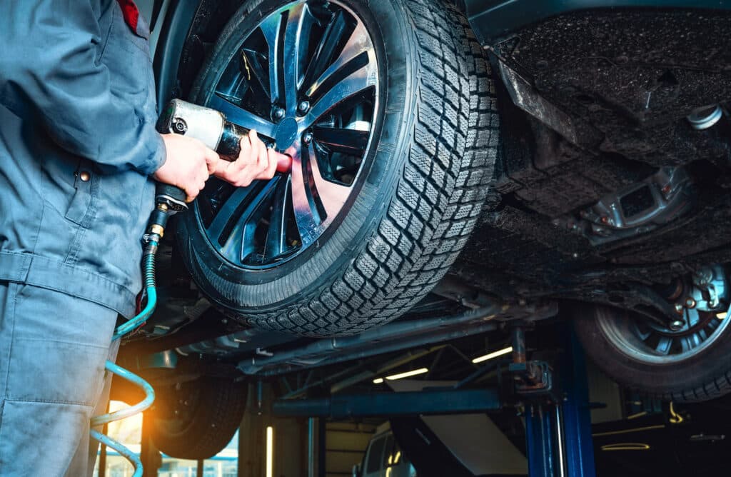 Ein Mechaniker verwendet ein pneumatisches Werkzeug, um Radmuttern an einem Autorad zu befestigen. Das Fahrzeug steht auf einer Hebebühne in einer Autowerkstatt, wo der Mechaniker im dunklen Arbeitsoverall seinen automatisch gespeicherten Entwurf der Reparaturen ausführt.